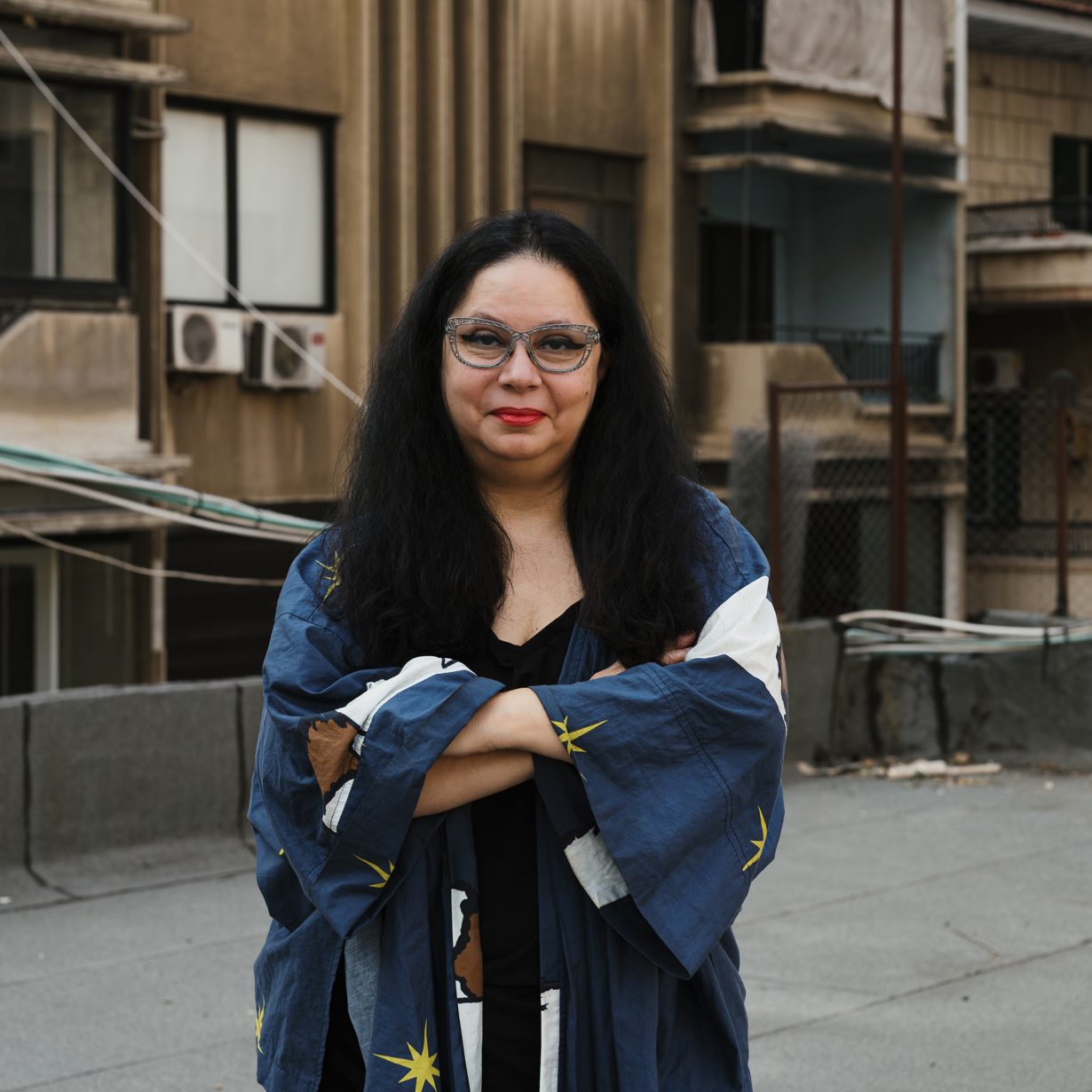 Portrait of curator Christine Tohmé. She is a 61-year old woman with black shoulder-length hair and wearing a blue dress. She is pictured  in front of some concrete buildings.