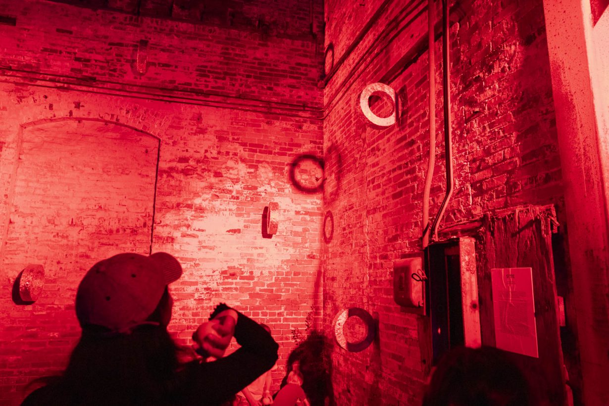 In a room lit by red light, a person wearing a cap seen from the back throws a ball at some rings attached to a brick wall.