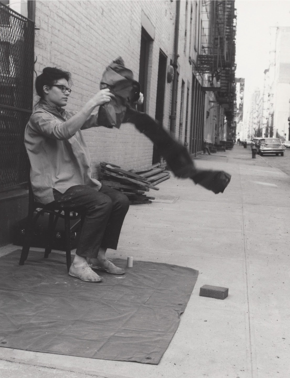 A black and white photograph of Alison Knowles sitting on a chair placed on a tarp in the street, and unfolding another tarp with both hands