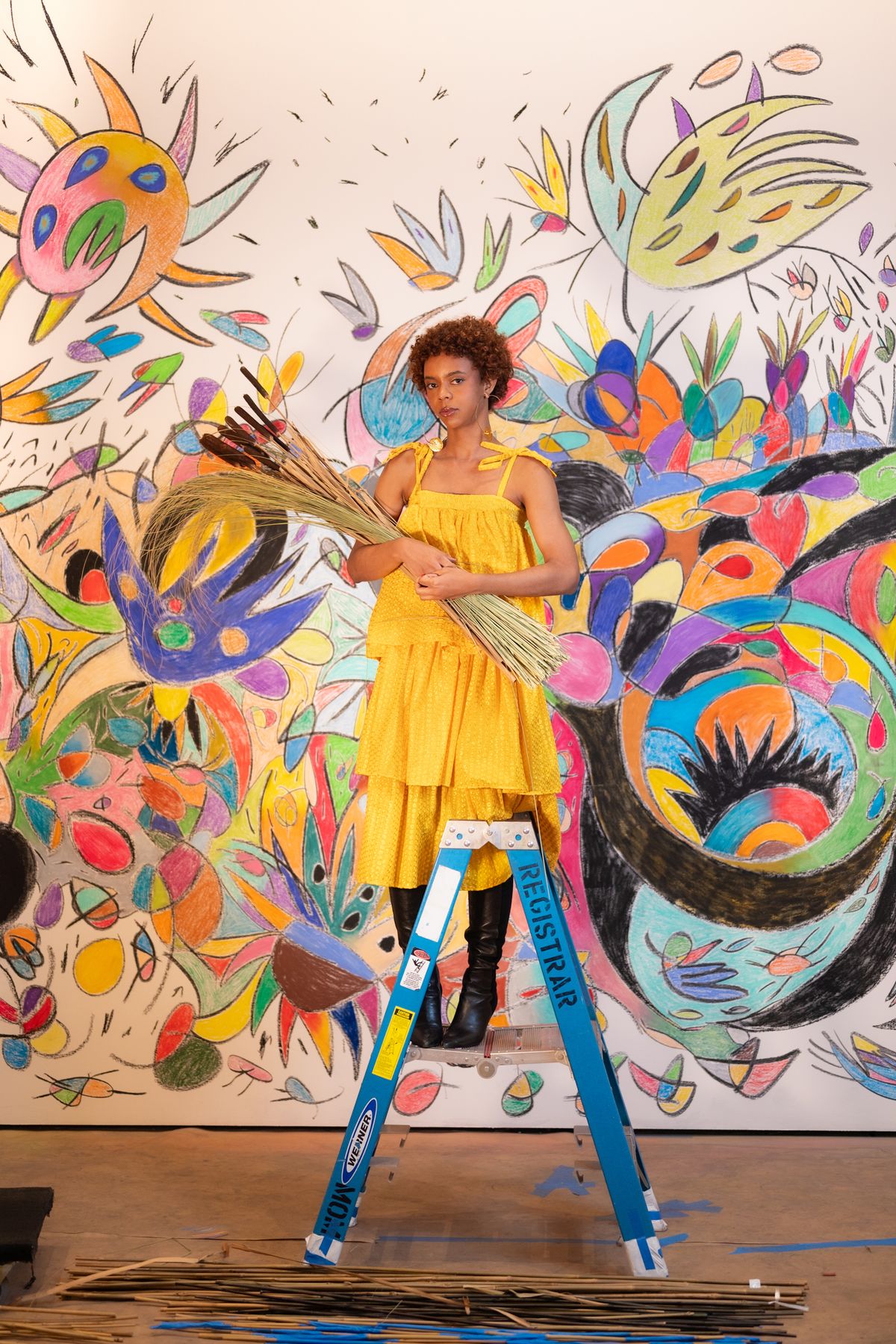 Tadáskía stands on a stepstool in front of a colourful mural. She is holding a bouquet of taboa, a reed-like plant.