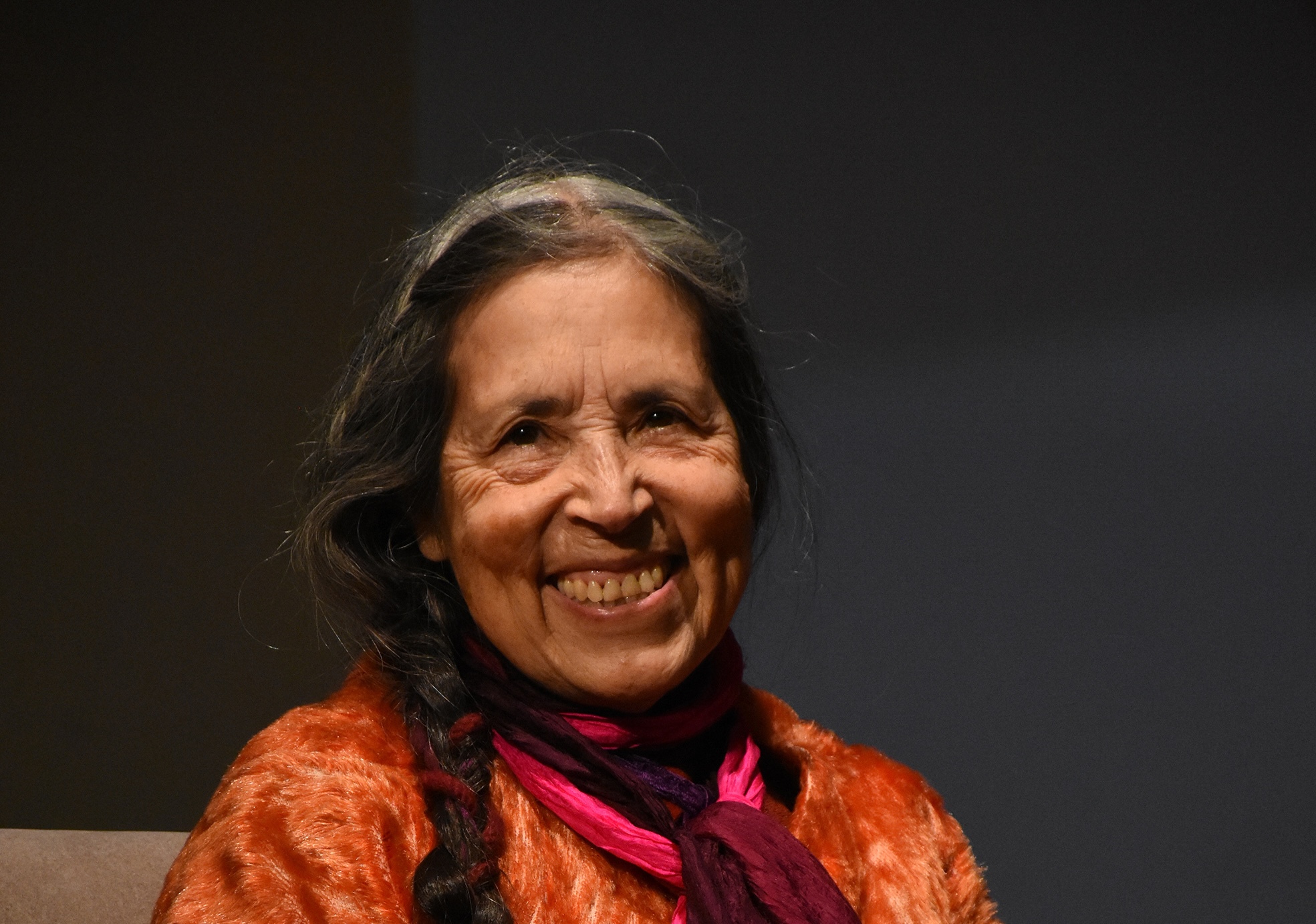 Cecilia Vicuña sitting and smiling, wearing an orange fur cardigan