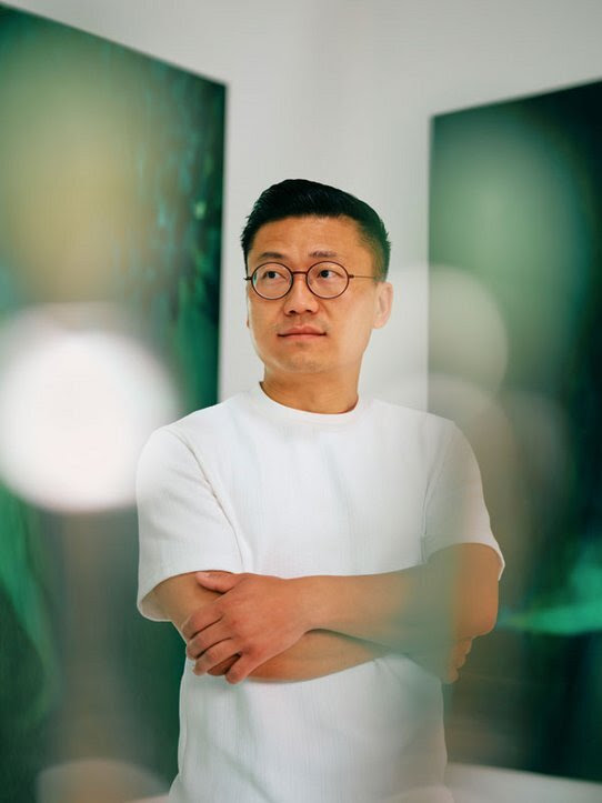 Xie Lei stands with his arms crossed in front of a blurred background containing paintings