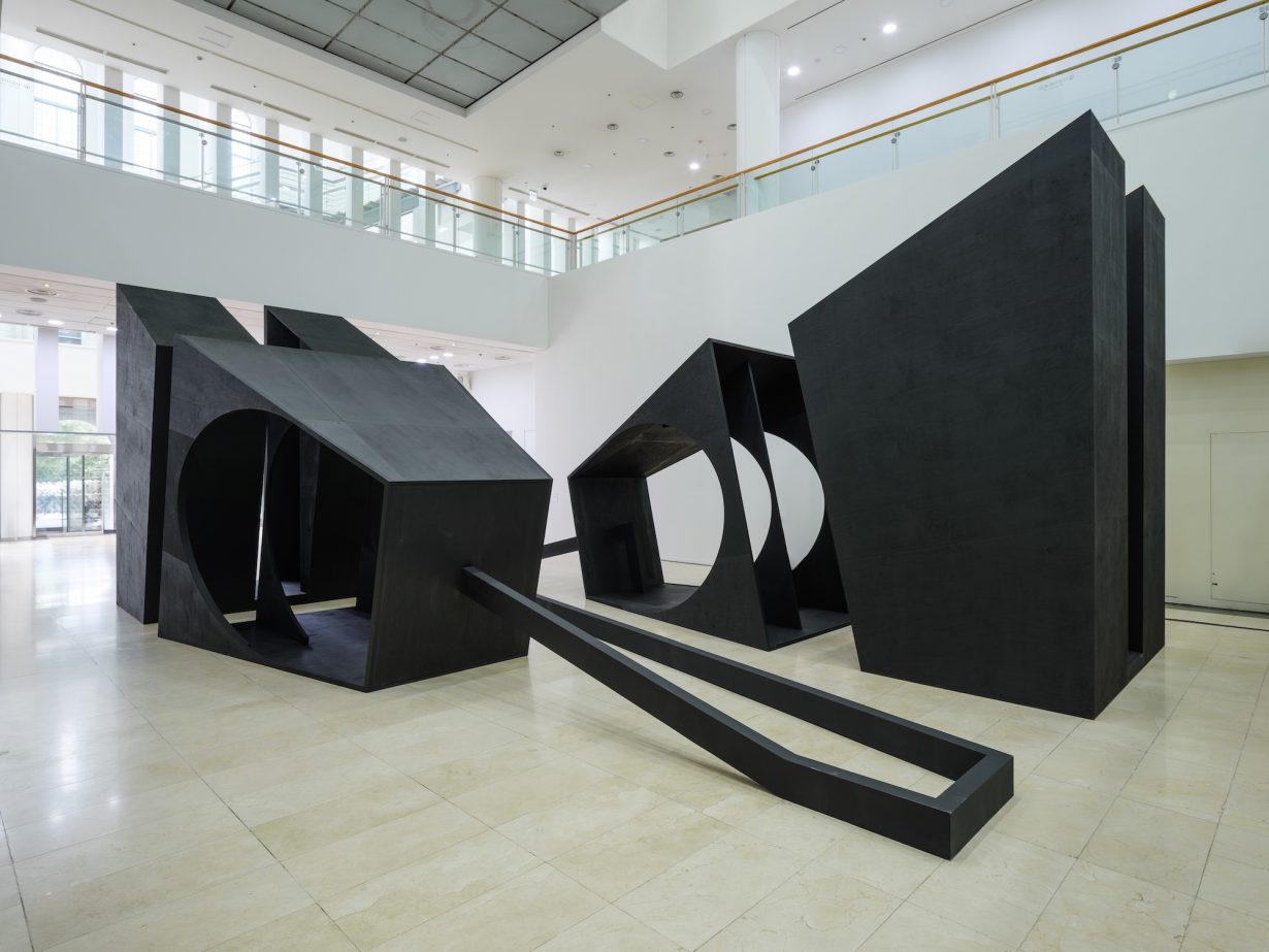 A largescale sculpture composed of four uneven hollow wooden black blocks with holes and connected by black wooden beams along the floor