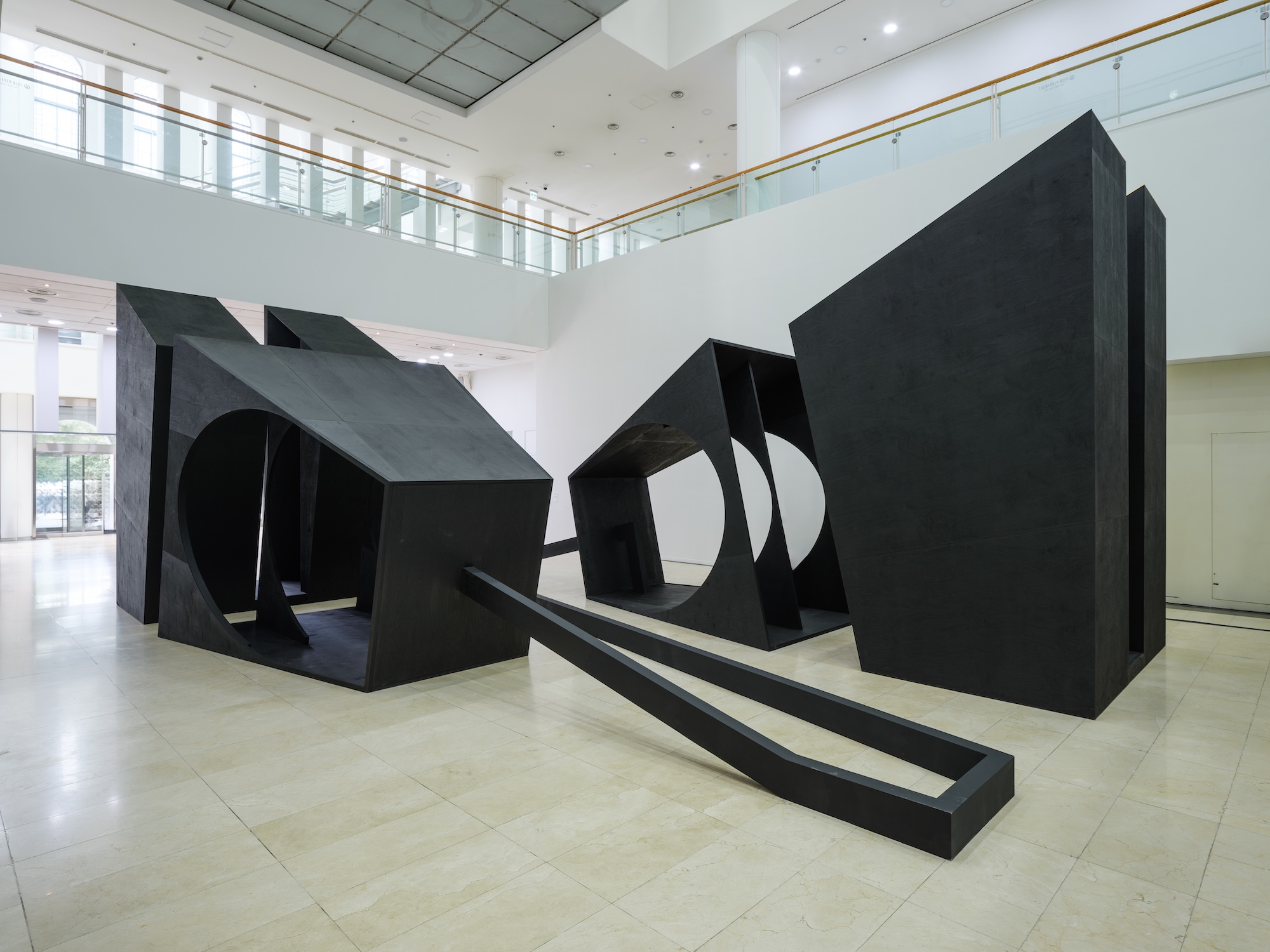 A largescale sculpture composed of four uneven hollow wooden black blocks with holes and connected by black wooden beams along the floor