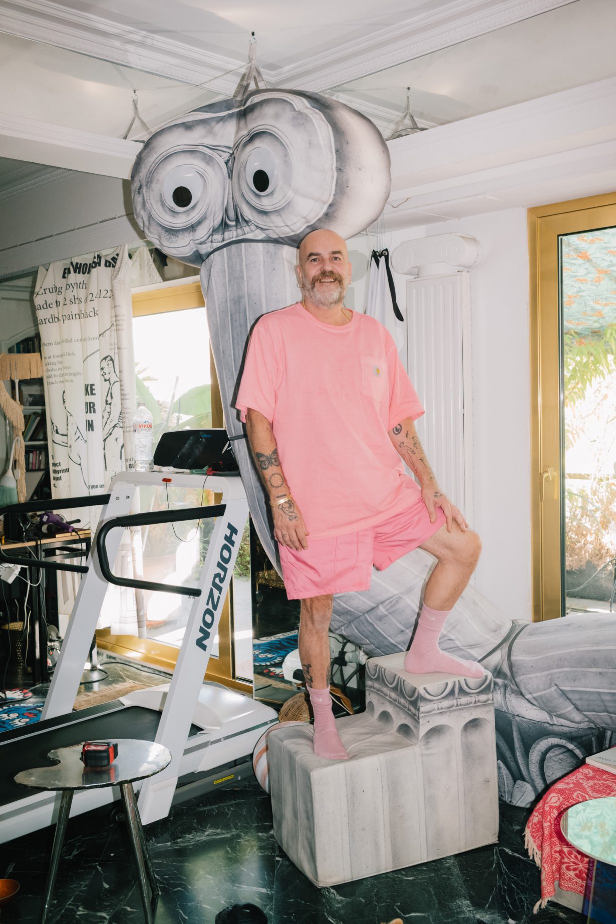 Andreas Angelidakis dressed in pink stands in his studio on a fake carved marble block in front of a fake stuffed ionic column that has eyes in the scrolls, hanging from the ceiling