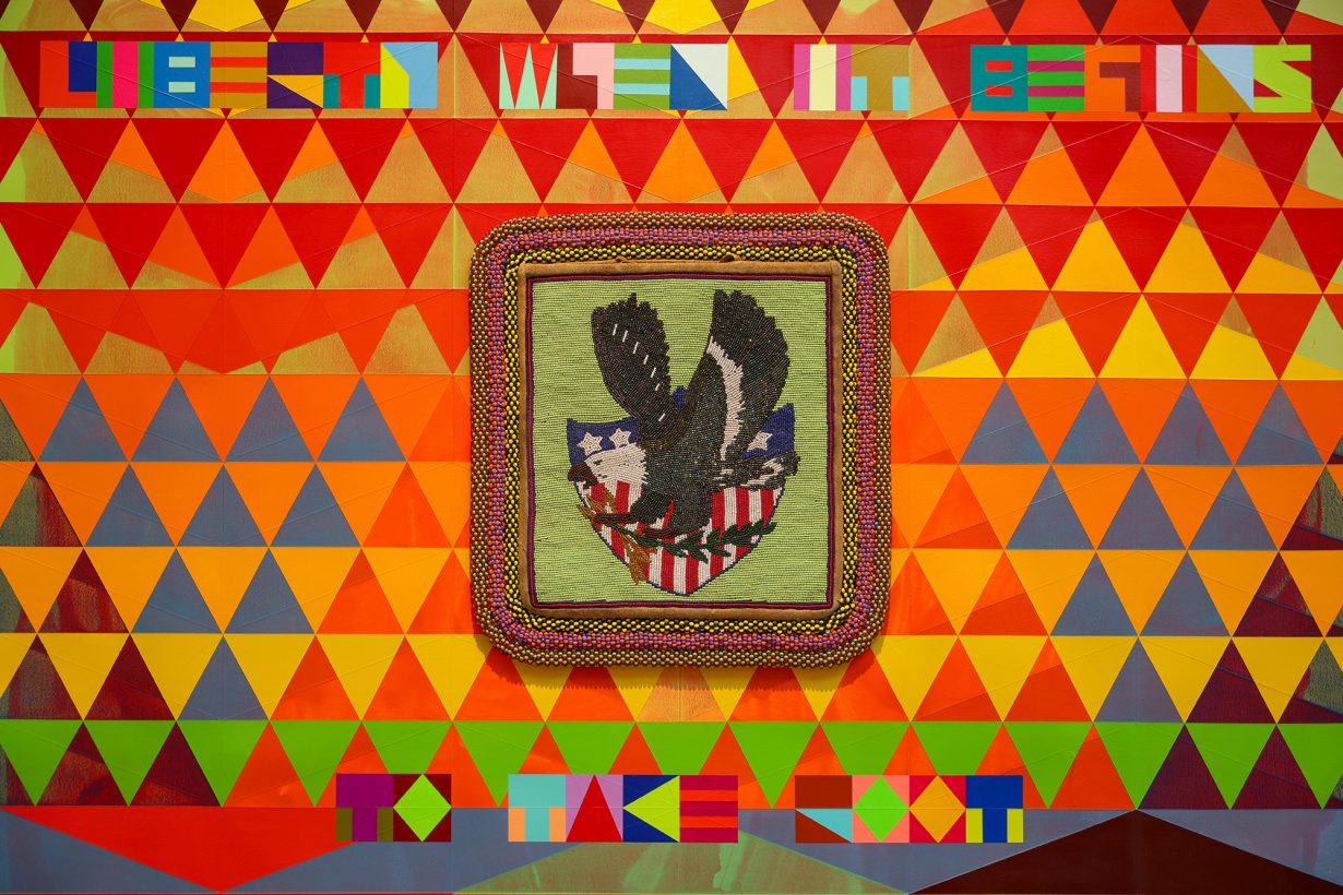 A wall decorated in colourful triangles with a framed and beaded image of an adaptation of the US seal