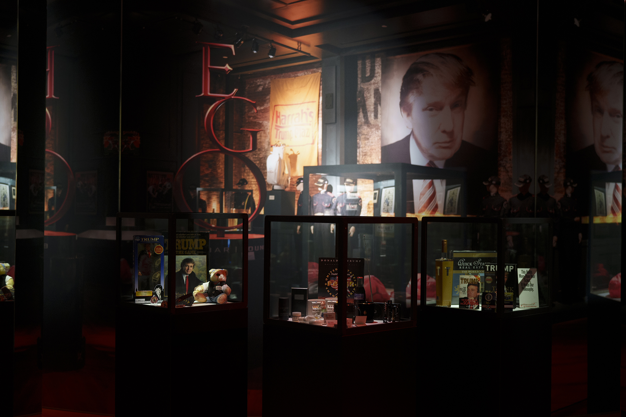 An exhibition view with images of Donald Trump on the wall, a red sculpture with the word 'Ego' and glass display cases filled with Donald Trump paraphernalia