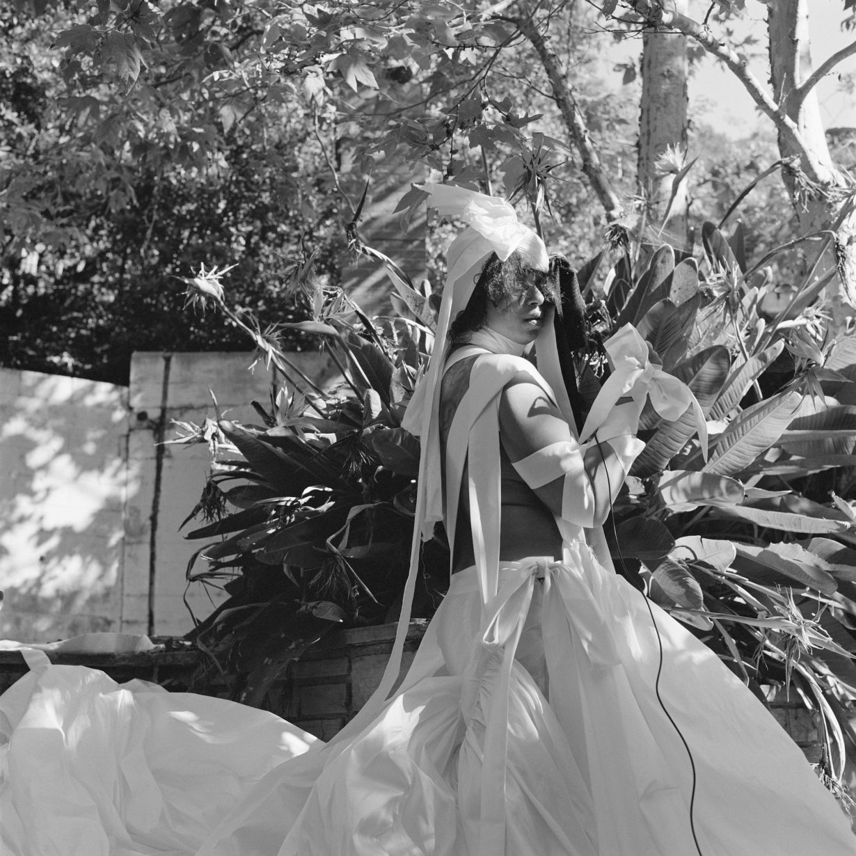 A black and white still of a person in a garden wearing a white costume