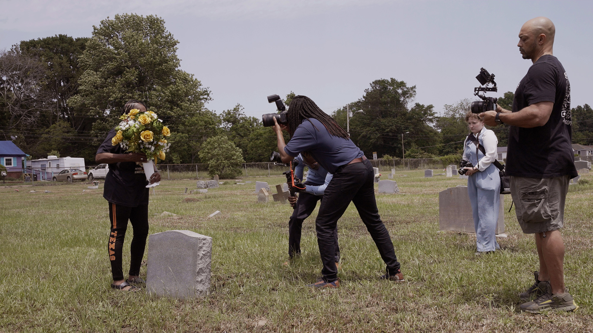 A film crew in a graveyard filming a person holding a bouquet of yellow flowers next to a tombstone