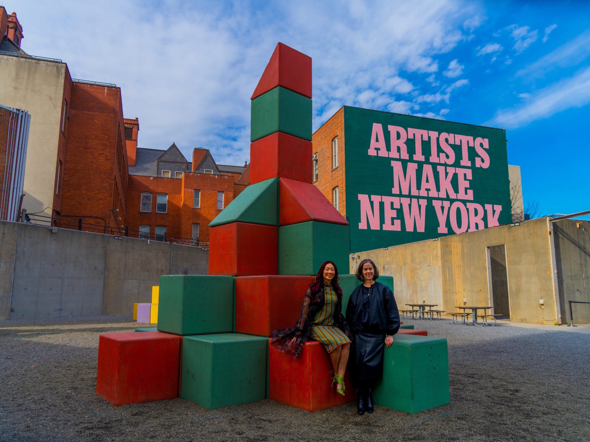 Sonya Yu and Connie Butler sitting on a green and red block sculpture by Yto Barrada in the MoMA PS1 courtyard. A mural on the museum facade in the background reads 'Artists Make New York'