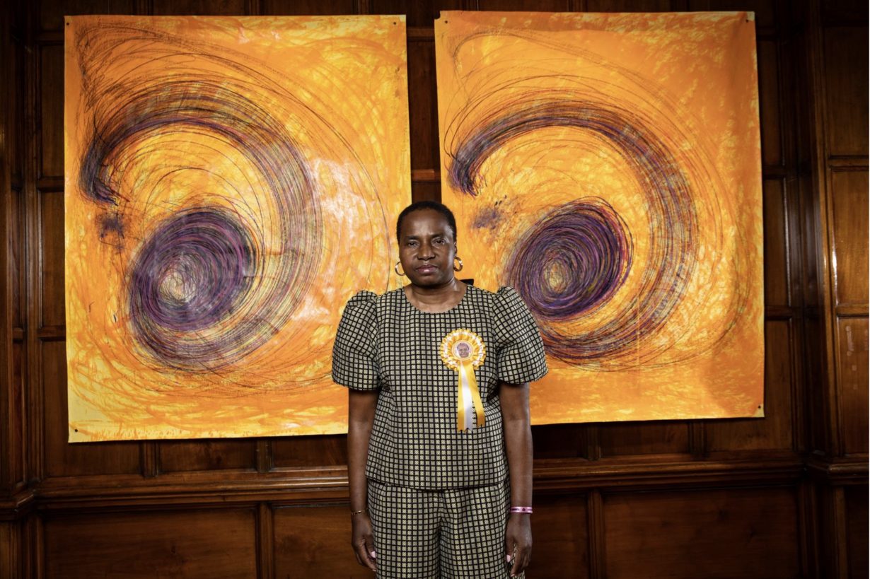 Nnena Kalu standing in front of two of her largescale, depicting large black and purple swirls on a yellow/orange background. The artist wears a yellow ribbon with her face on it.