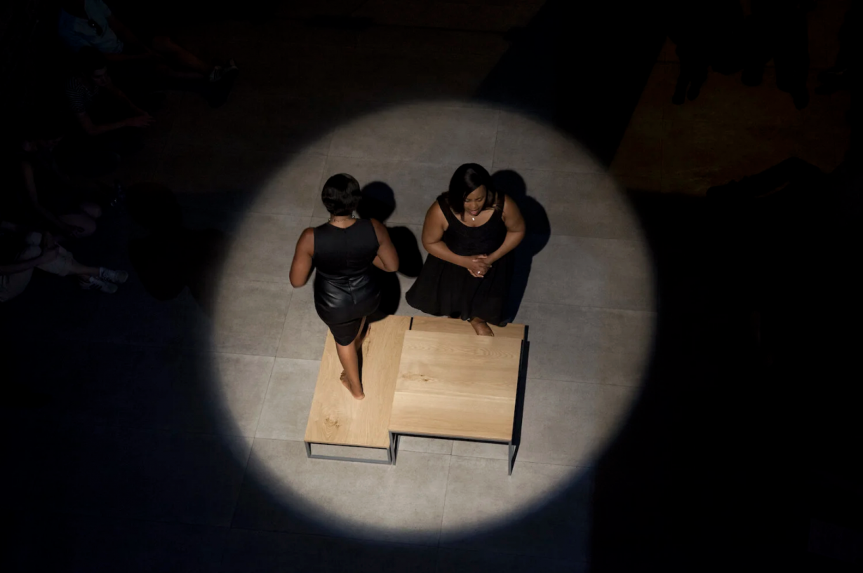 Gabrielle Goliath's Elegy - Noluvo Swelindawo, a performacne work. Image shows two women dressed in black standing on a spotlit stage. 