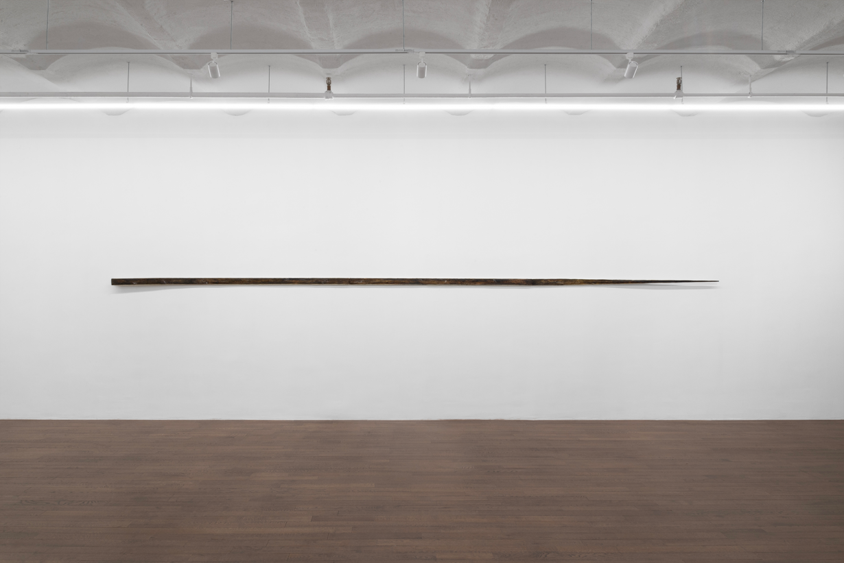 A long, thin, tapered sculpture on a white wall. 