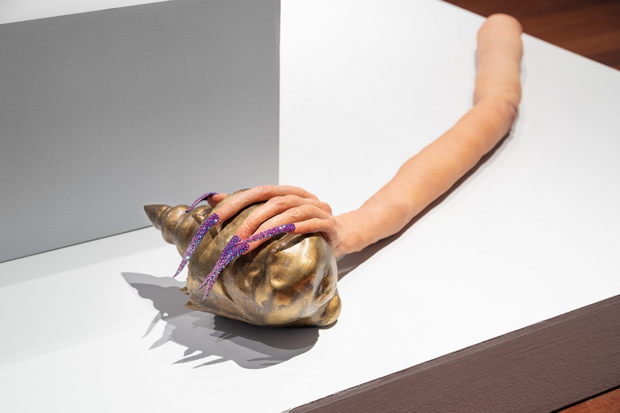 A sculpture resting on a white plinth of an elongated arm and hand with long, pointed, purple-studded nails is wrapped around a bronze budda head