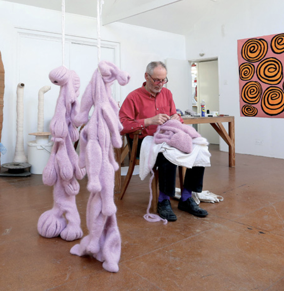 Nicholas Pope sat stitching in his studio surrounded by works. He is wearing a red shirt and glasses. 