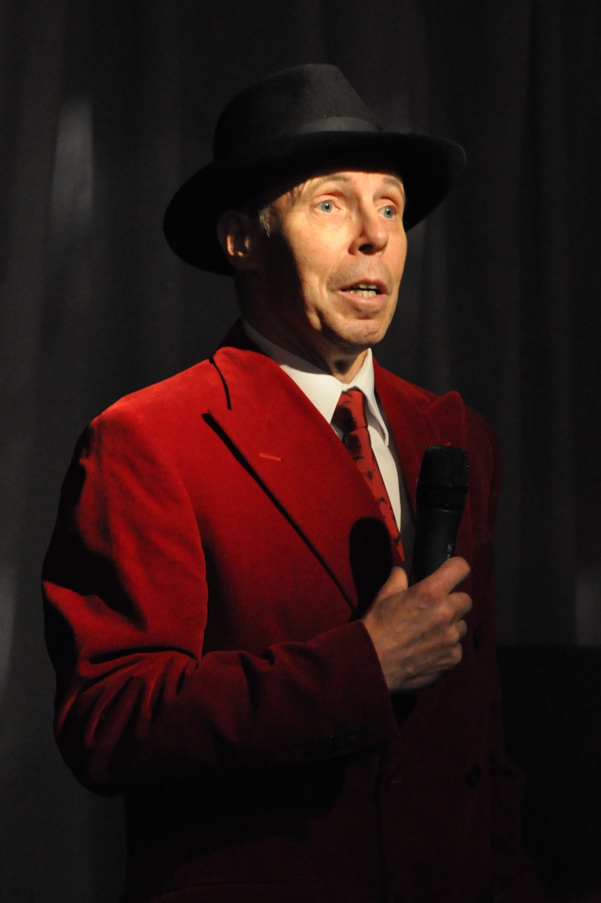 Gunther Von Hagens in a red suit and black hat speaking into a mircrophone