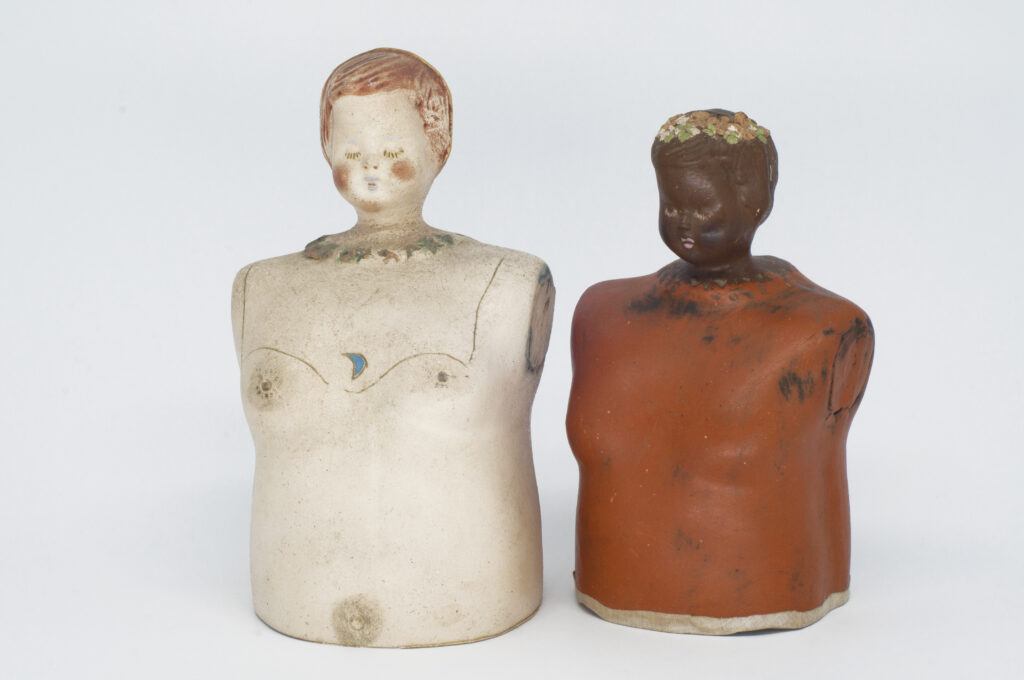 Photo of two works by Marlies Ritter, featuring ceramic dolls' heads on oversized torsos. One cream, one brown. 