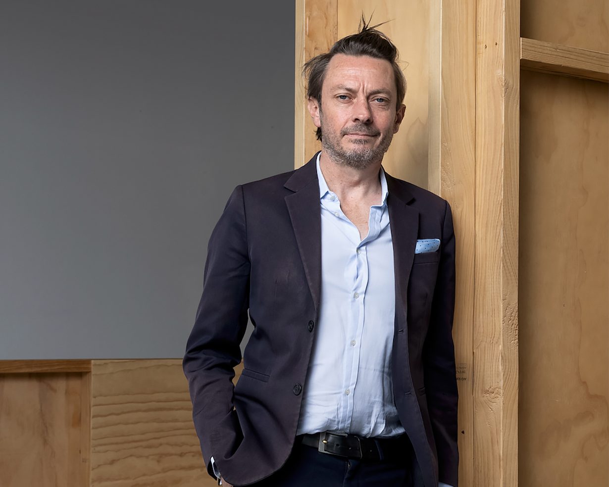 Photo of Mark Feary standing up in a navy suit, light blue shirt, no tie but a picket handkerchief.