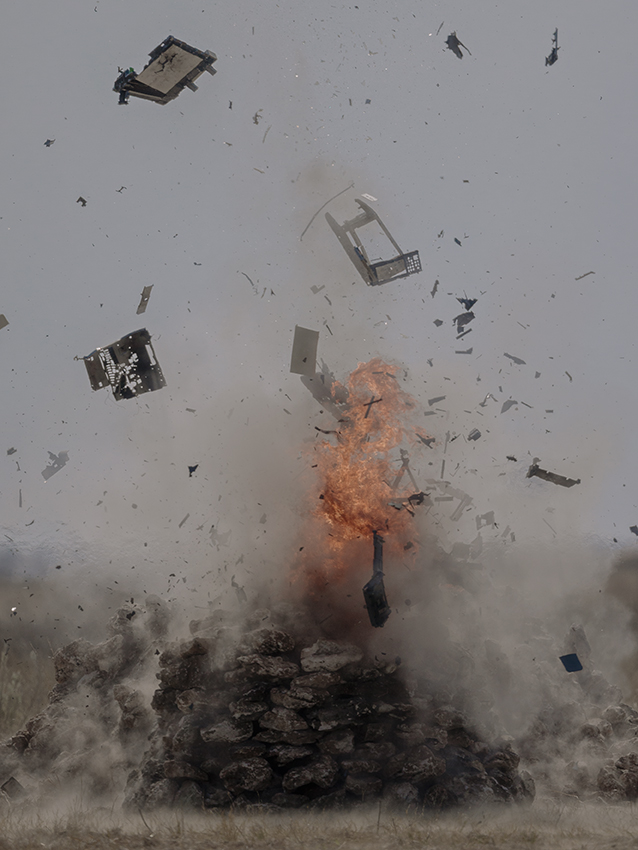 Colour film still of an explosion with debris flying in the air. In the centre a fire burns.